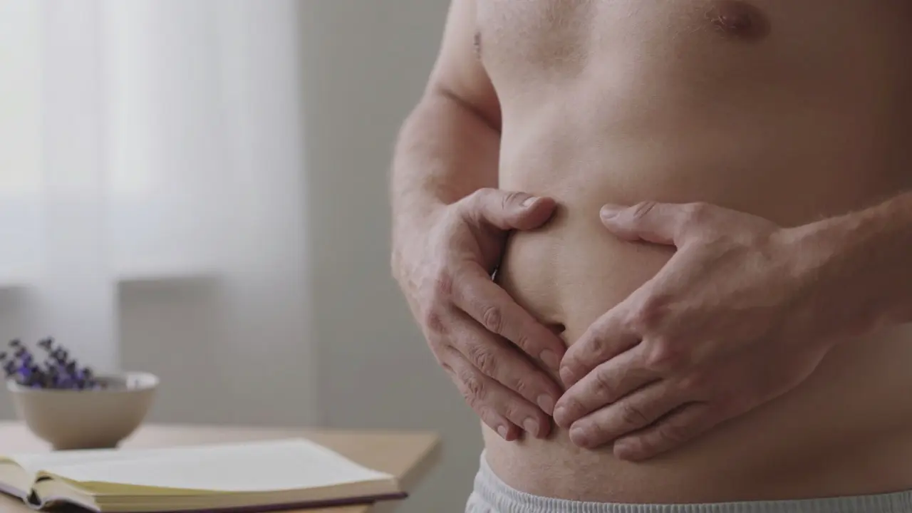 Muž se dotýká svého břicha doma v klidném ranním světle, pokojný výraz, bez nátlaku.